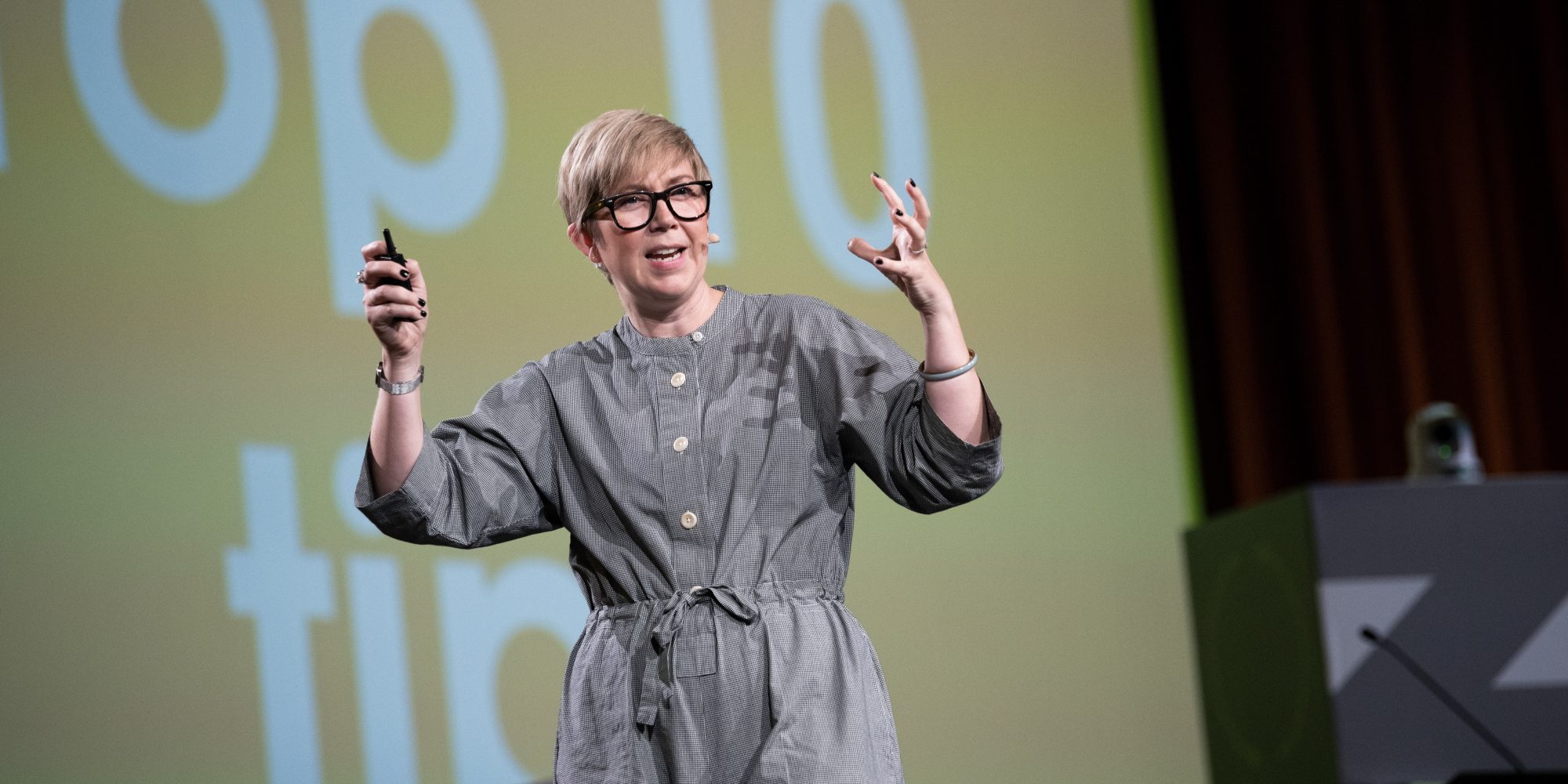 Katie speaking on stage at an international zero waste conference, wearing a grey shirt, jumpsuit, back glasses and hand expressions