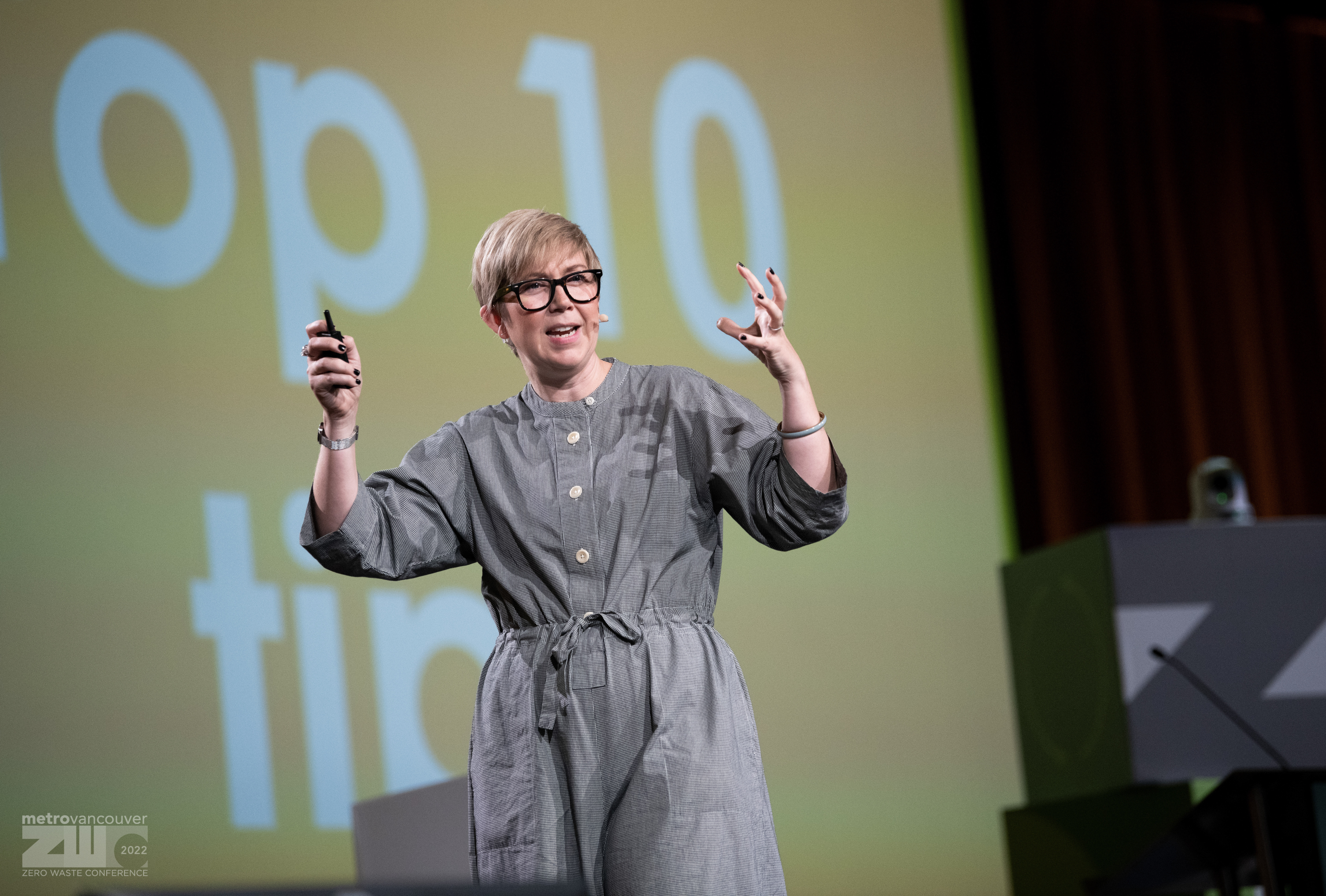 Katie speaking on stage at an international zero waste conference, wearing a grey shirt, jumpsuit, back glasses and hand expressions